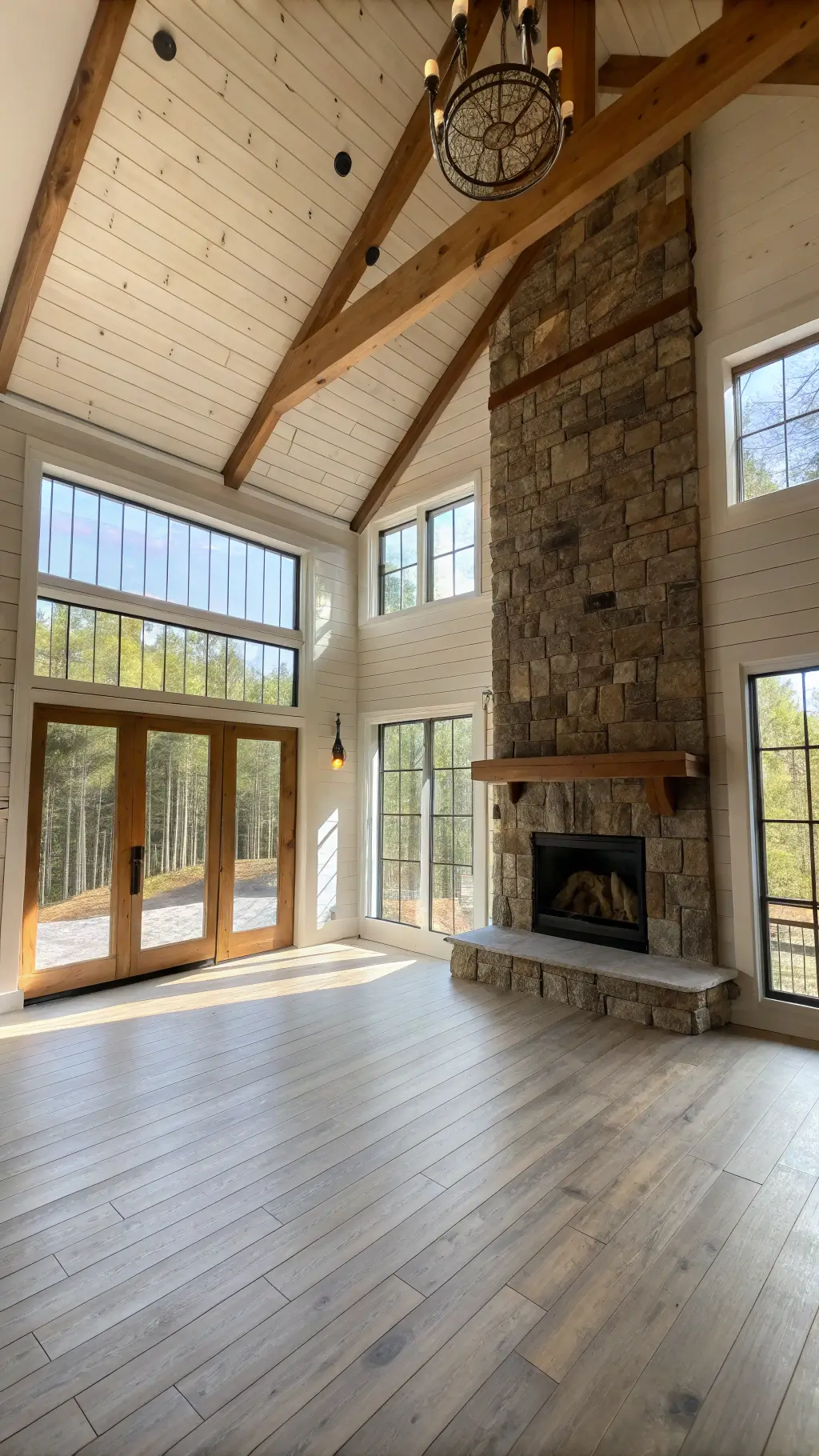 Image of a cozy 400 sq ft cabin interior with morning light streaming through large windows, featuring an open-concept living area. The space has a 12-foot high exposed pine beam ceiling, whitewashed plank walls, and a stone veneer fireplace. Aged bronze sliding barn door leads to the bedroom, while reclaimed wood floating shelves adorn the walls.