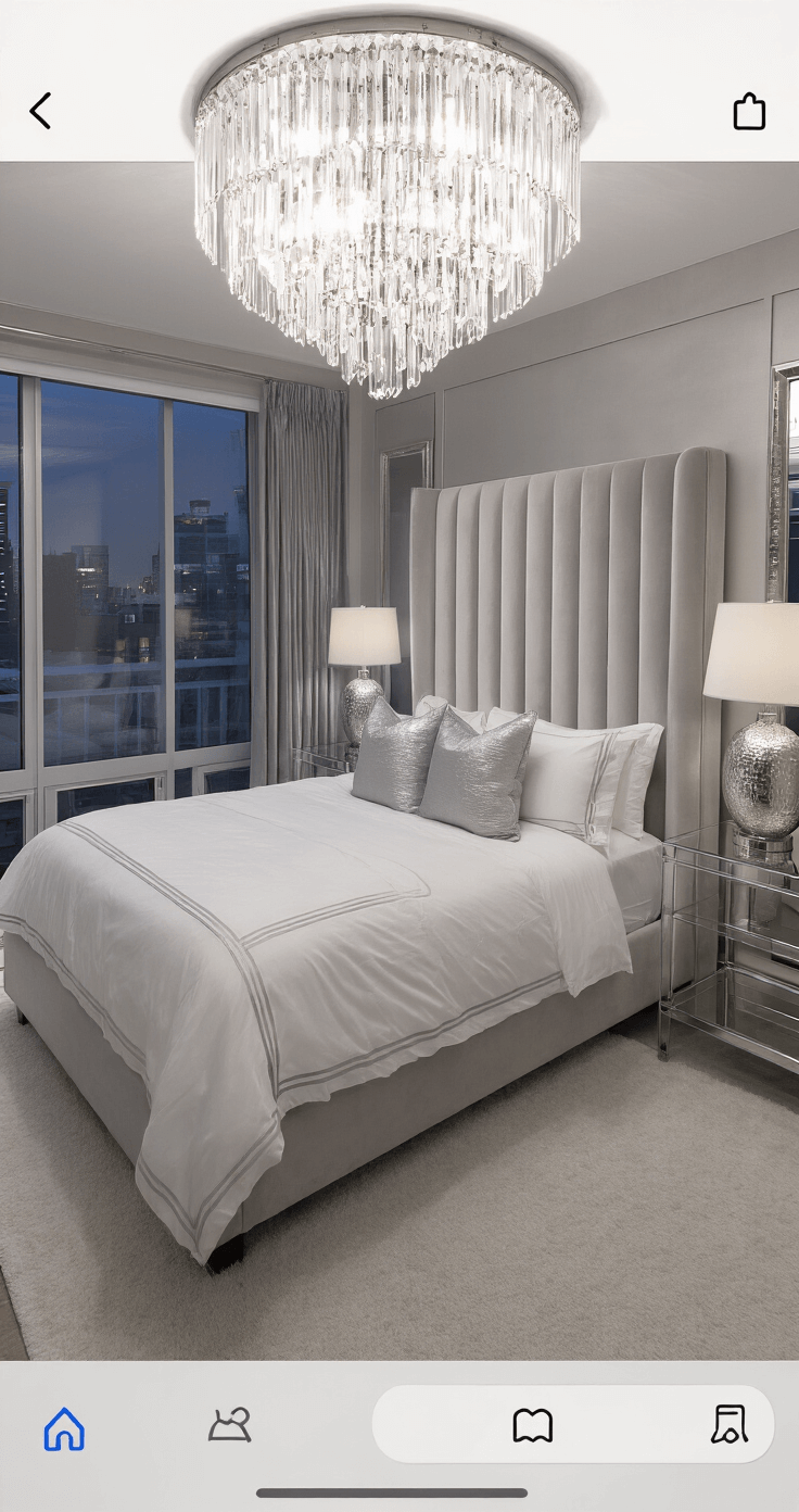 Luxe hotel-style bedroom at twilight featuring a king bed with a channeled pearl grey velvet headboard, white bedding, silver accent pillows, and a dimmed crystal chandelier, with floor-to-ceiling mirrors, lucite side tables, and plush white carpet in a monochromatic grey and silver palette.