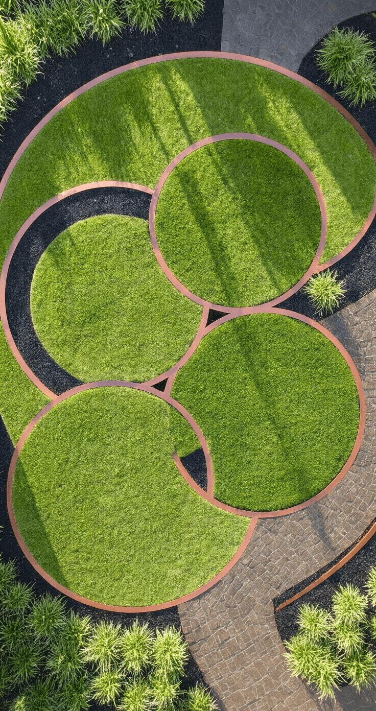 Overhead view of three interlocking circular lawns with 12' diameters, showcasing contemporary garden design with morning dew on fresh-cut grass, copper edging, and black mondo grass borders, highlighted by geometric mowing patterns and dramatic shadows from the rising sun.