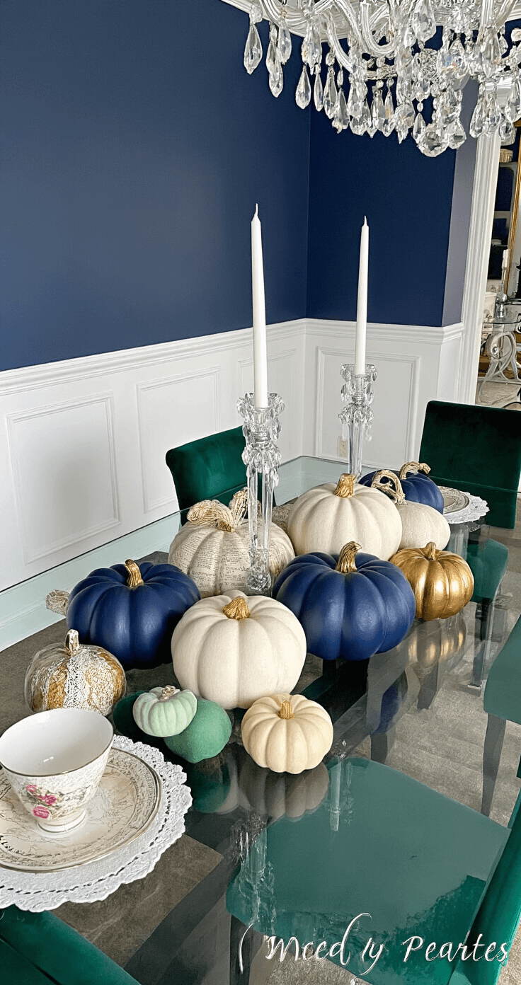 An elegant dining room featuring deep navy walls and white wainscoting, centered around a modern glass-top dining table surrounded by emerald green velvet chairs. The table displays a colorful collection of foam pumpkins in matte white, glossy navy blue, and metallic gold leaf, some adorned with lace patterns and book pages. Crystal candlesticks with white tapers accentuate the centerpiece, illuminated by a soft glow from an overhead crystal chandelier.