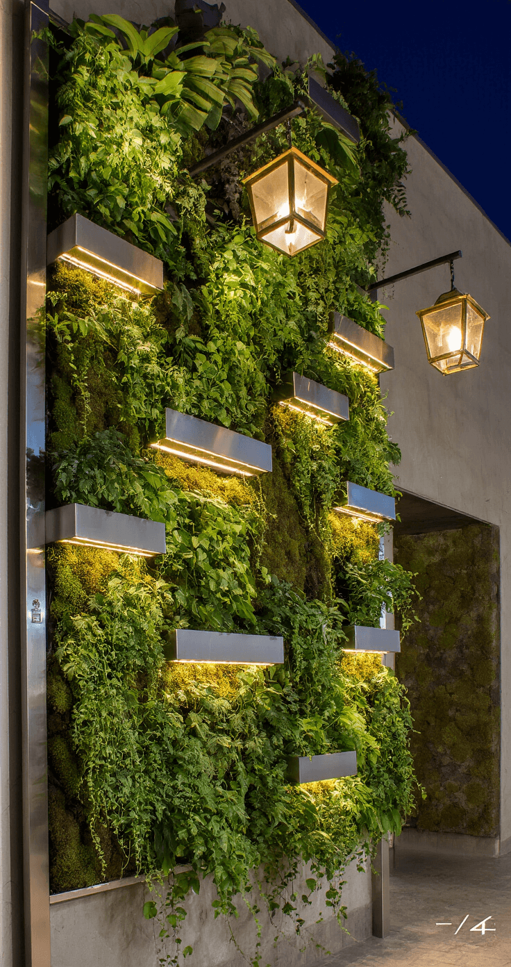 Vertical garden wall at night featuring stainless steel sconces, preserved moss panels, and trailing vines, enhanced by hidden LED strips for dramatic lighting, complemented by suspended aged brass lanterns.