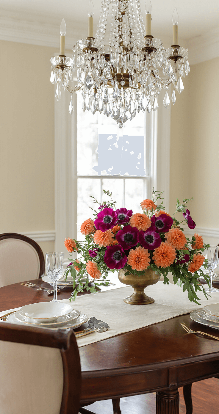 Elegant dining room with winter floral arrangement of bold magenta anemones and warm clementine orange dahlias in a vintage brass vessel on a mahogany table, featuring crown molding and a crystal chandelier. Cream silk table runner and bone china place settings enhance the sophisticated color palette against neutral walls and rich wood tones.
